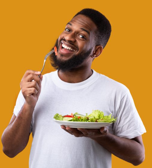 Cheerful Black Guy Eating Salad Posing Holding Plate And Fork Enjoying Healthy Meal Standing Over Yellow Studio Background, Smiling To Camera. Vegan Dinner Recipes, Weight Loss And Male Nutrition
